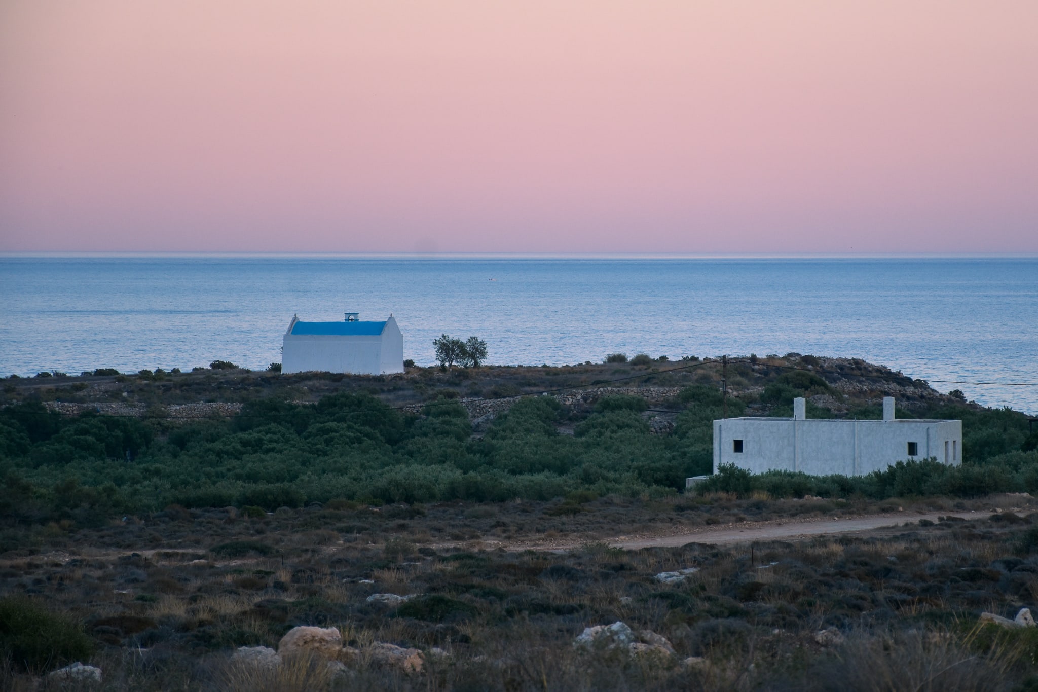 Sunset view from a plot in Xerokampos, east Crete. Sky in the background has a bit of violet color, sea is calm and blue and on the landscape horizon there is a small church of Saint Antonios. It has a white walls and a typical greek blue roof, which makes a perfect contrast with the sea.