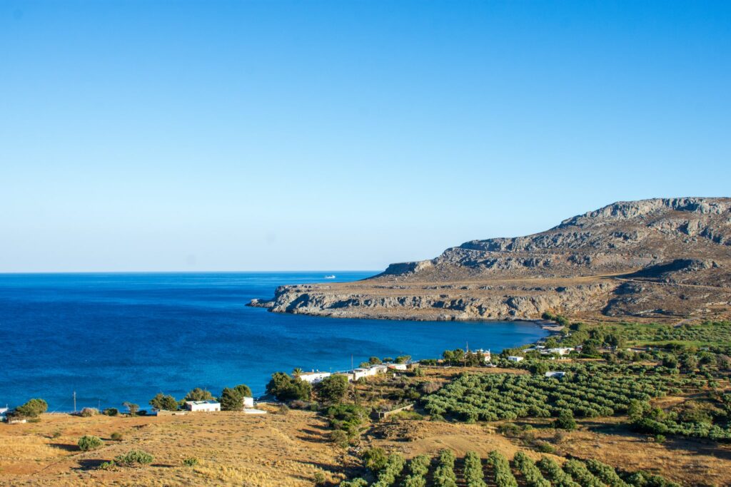 View at the bay of Kato Zakros. There is a sea with a deep dark blue color on the left and on the right some green olive trees. In the background there is a big mountain with a road to Zakros.